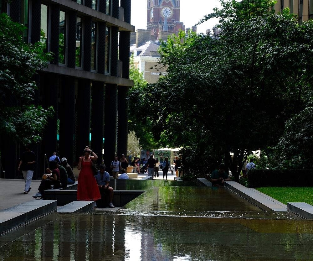 6 View along a shallow urban water feature with people sitting under trees; the distant clocktower of St Pancras reflected in the pool.