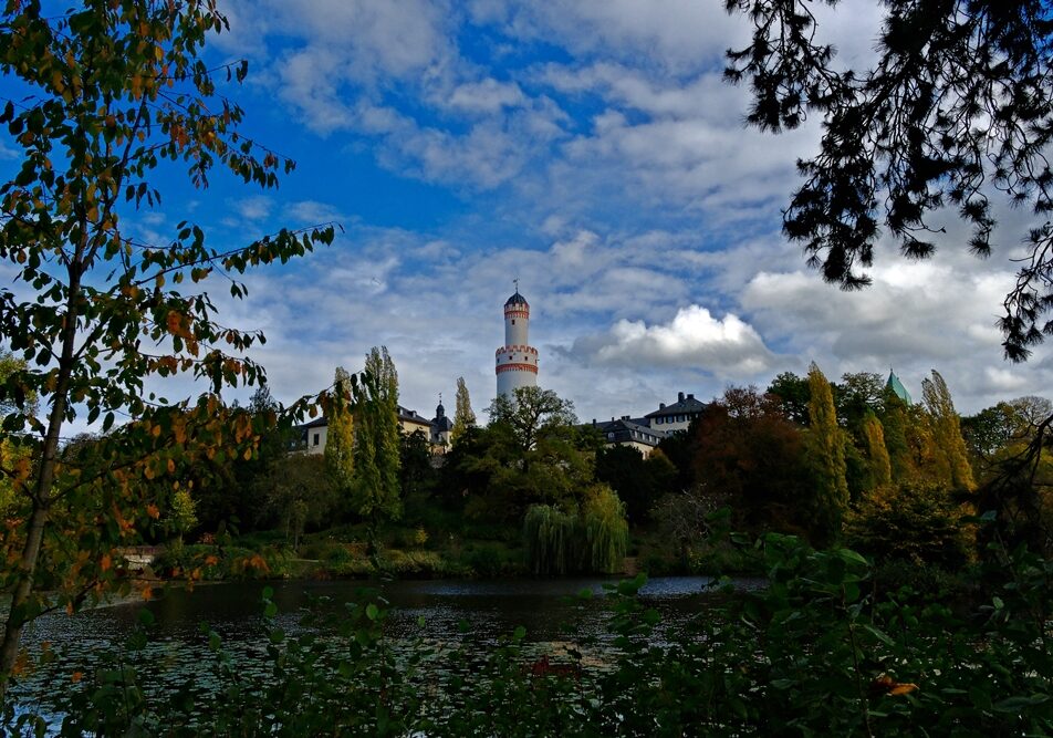 12 months project_BH_15 October 2025 A scenic view of a historic castle with a tall white tower featuring red accents, surrounded by lush green and autumn-colored trees, reflected in a calm lake under a partly cloudy blue sky.